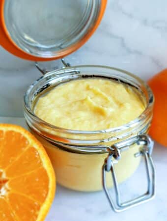 An open jar of orange curd next to half an orange.