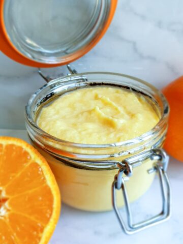 An open jar of orange curd next to half an orange.