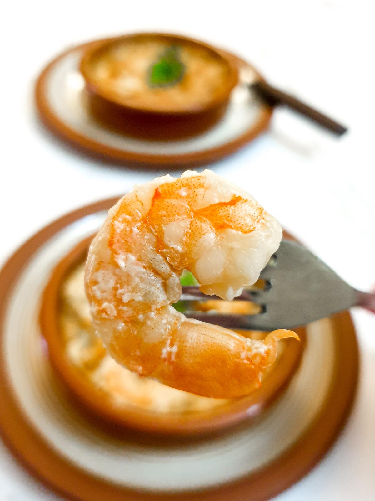 Close up of a prawn on a fork held over a dish of prawn and scallop au gratin.