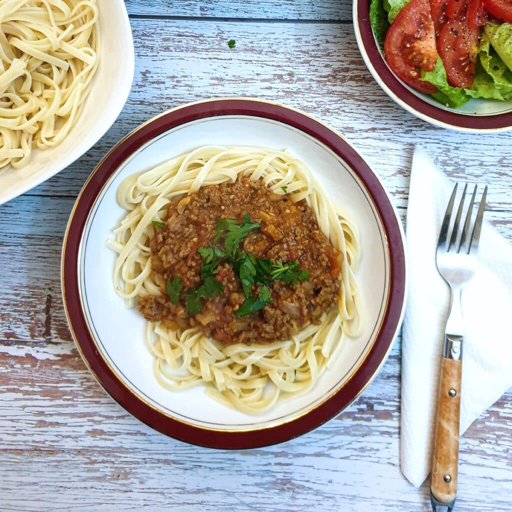 Bolognese sauce on a bed of linguine on a table next to a bowl of green salad.