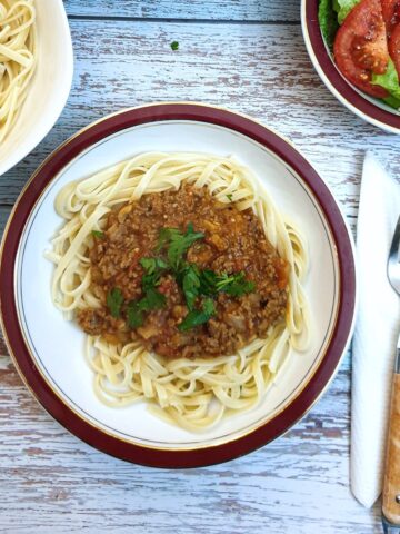 Bolognese sauce on a bed of linguine on a table next to a bowl of green salad.