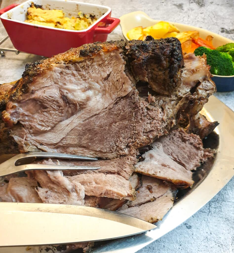 A leg of lamb on a table with dishes of vegetables.