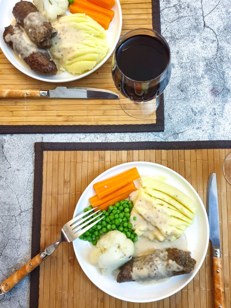 Two plates of steak roulade with vegetables and mashed potatoes, covered in horseradish gravy.