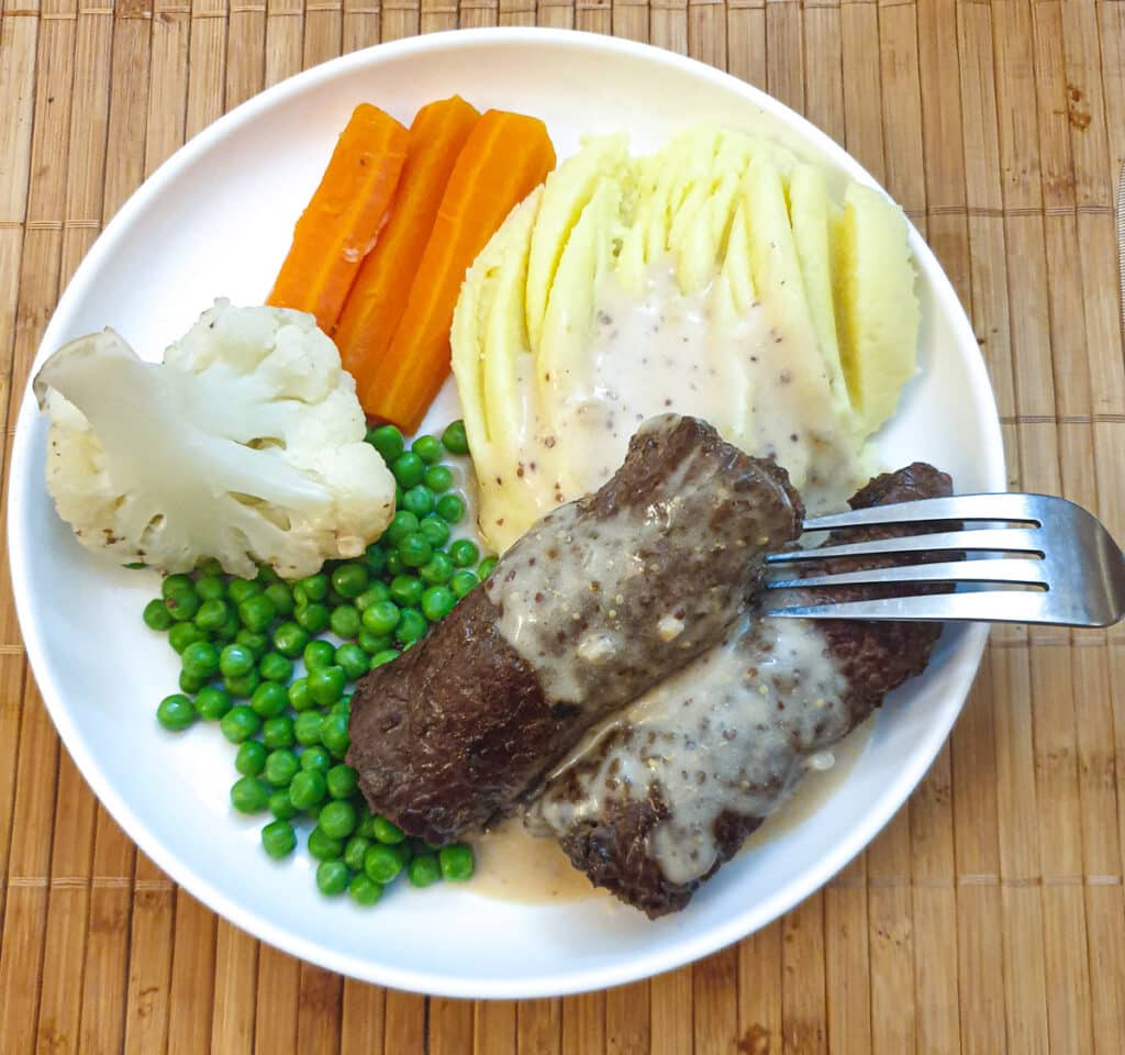 A white dinner plate with vegetables and two steak roulades, drizzled with gravy.