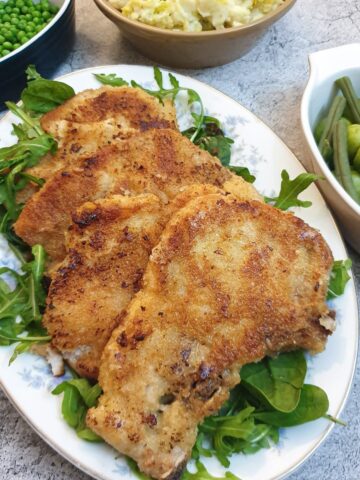 4 crispy crumbed pork chops on a platter next to dishes containing vegetables.