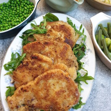 4 crispy crumbed pork chops on a platter next to dishes containing sauce and vegetables.