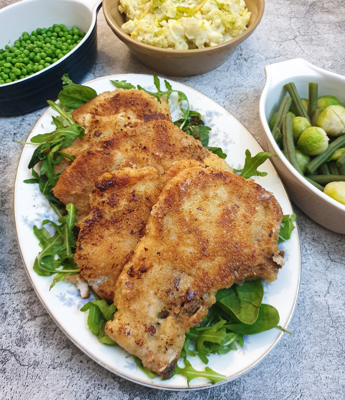 4 crispy crumbed pork chops on a platter next to dishes containing vegetables.