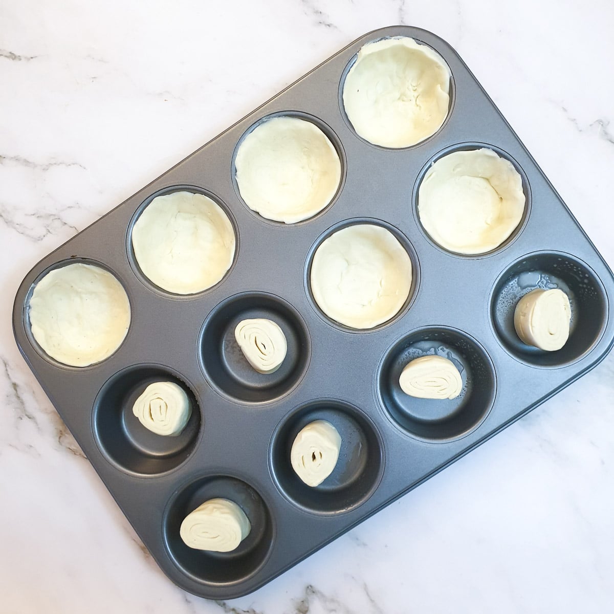 A 12-hole muffin tin being lined with pastry.