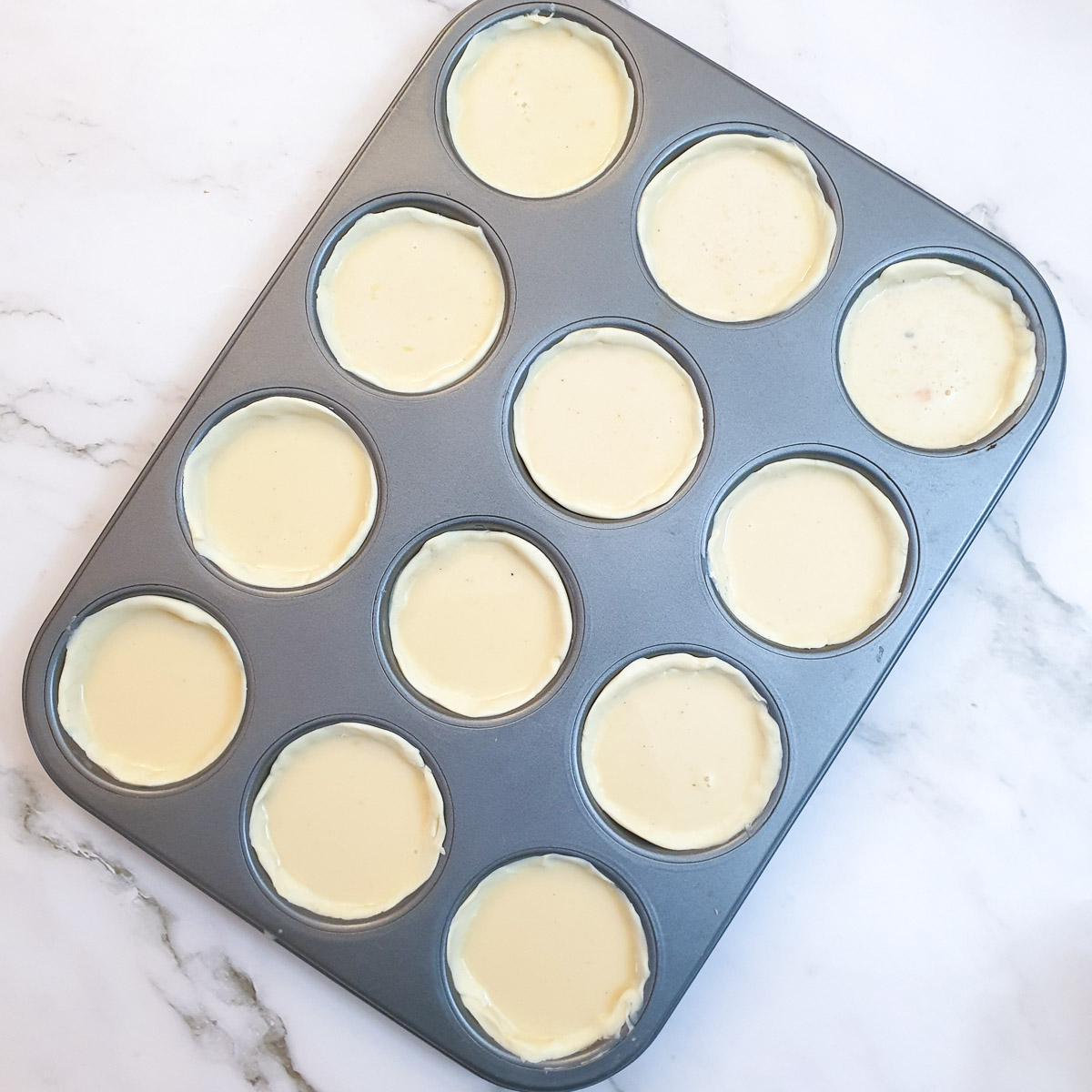 A 12-hole muffin pan containing 12 unbaked Portuguese custard tarts.