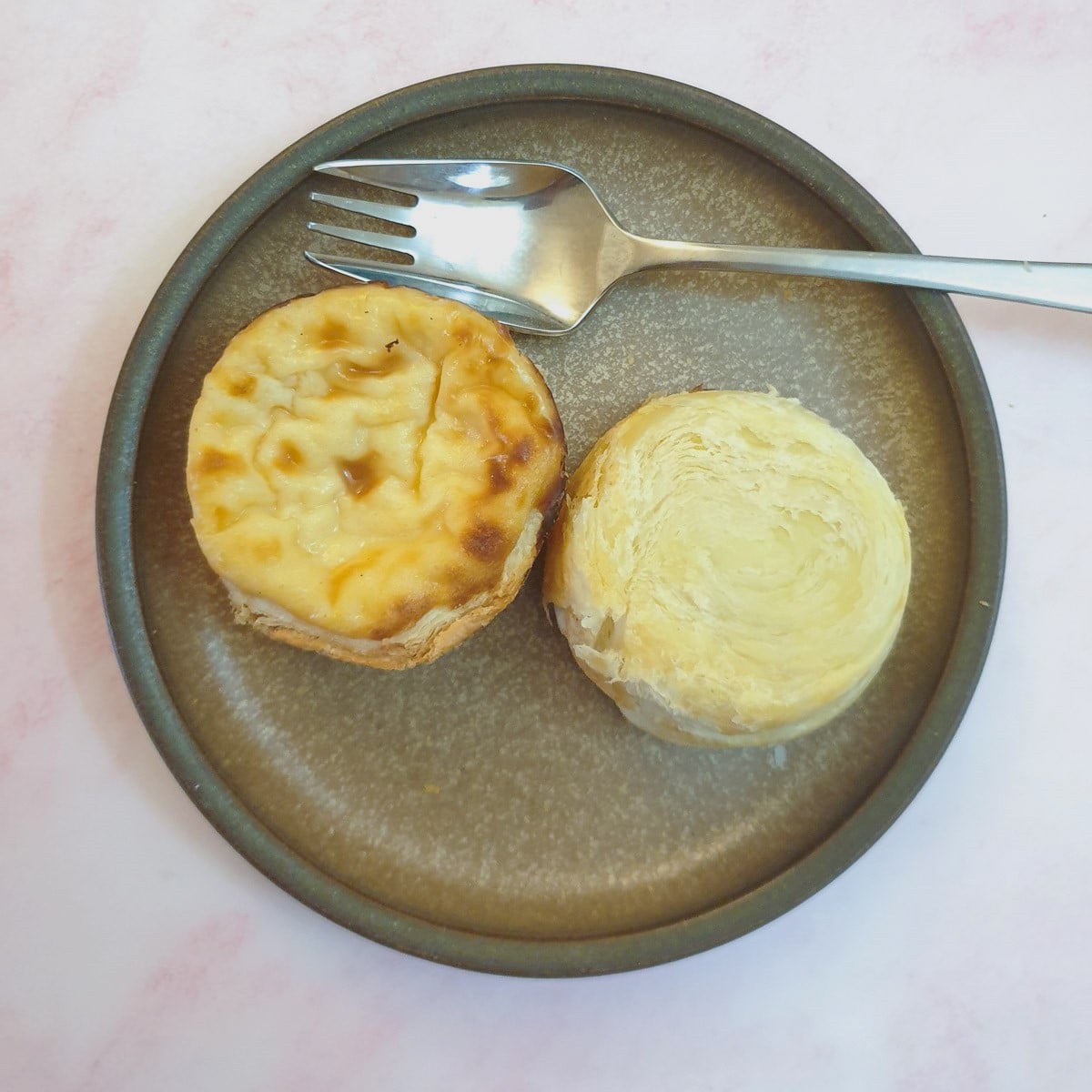 Two portuguese custard tarts on a plate, with one turned upside down to show the laminations in the pastry.