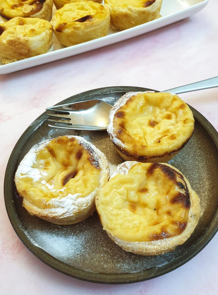 3 Portuguese custard tarts lightly dusted with icing sugar on a brown plate with a fork.