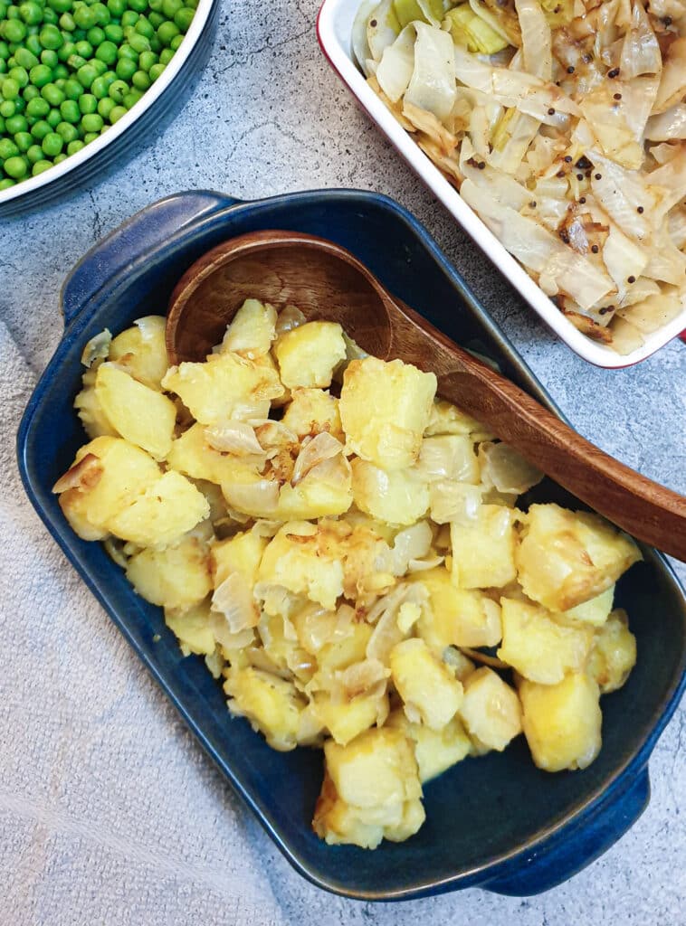 Saute potatoes and onions in a blue dish next to a dish of cabbage and a dish of peas.