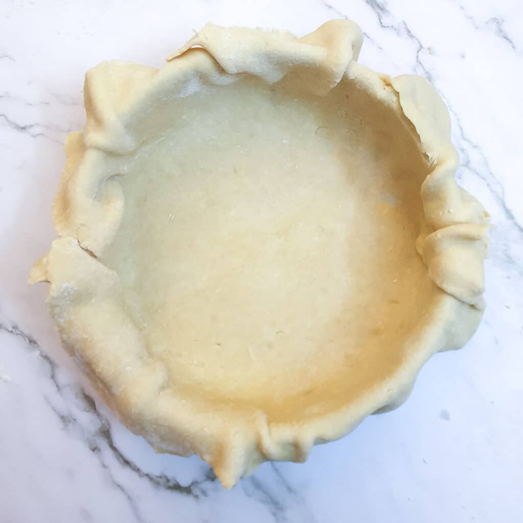 A round baking dish lined with pastry, with the pastry overhanging the edges of the dish.