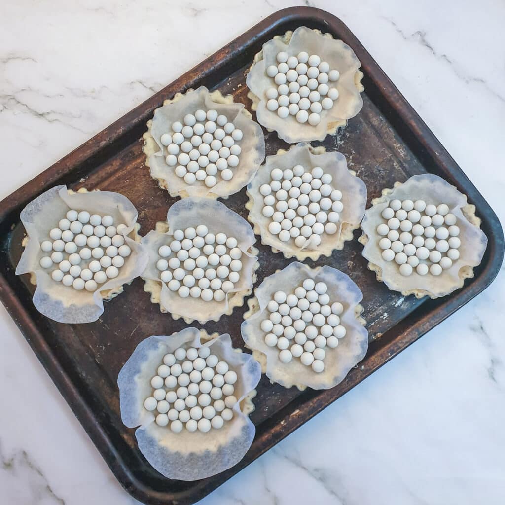 8 unbaked pastry shells lined with baking parchment and baking beans on a baking tray.