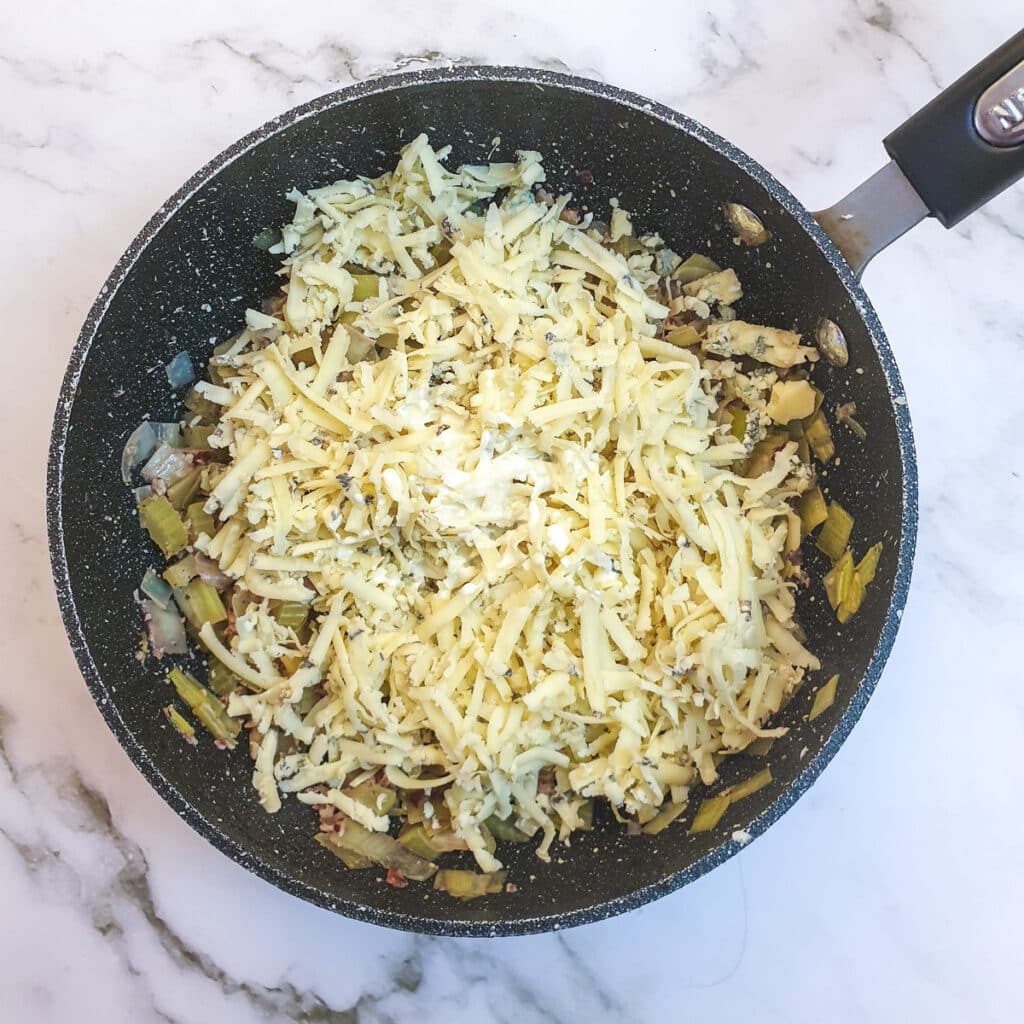 Grated Stilton cheese added to leeks and celery in a saucepan.