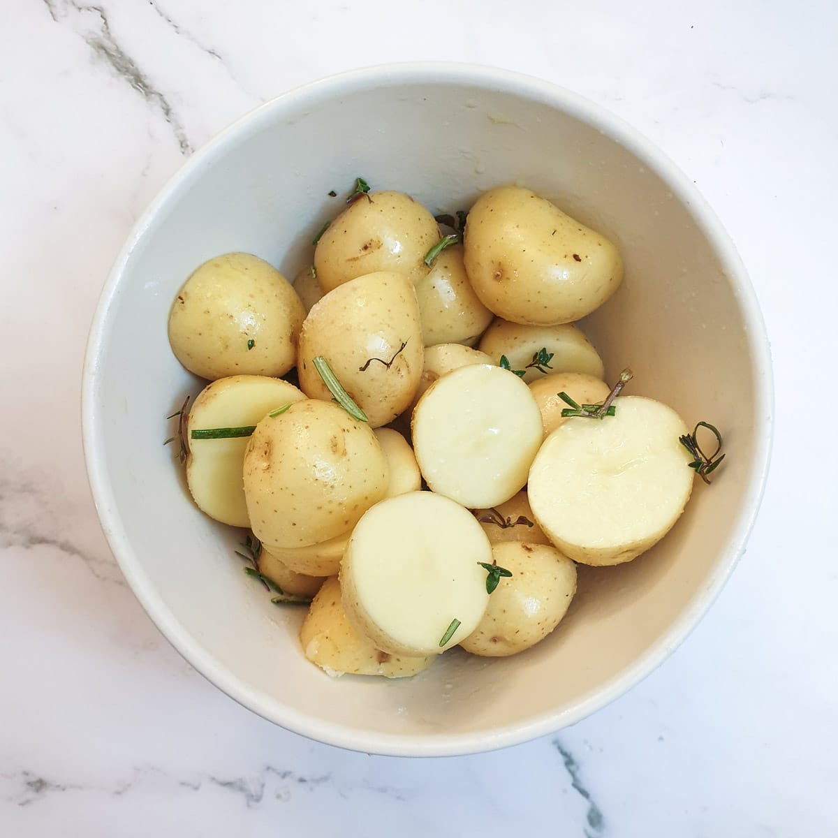 Halved baby potatoes in a bowl with olive oil, rosemary and thyme.