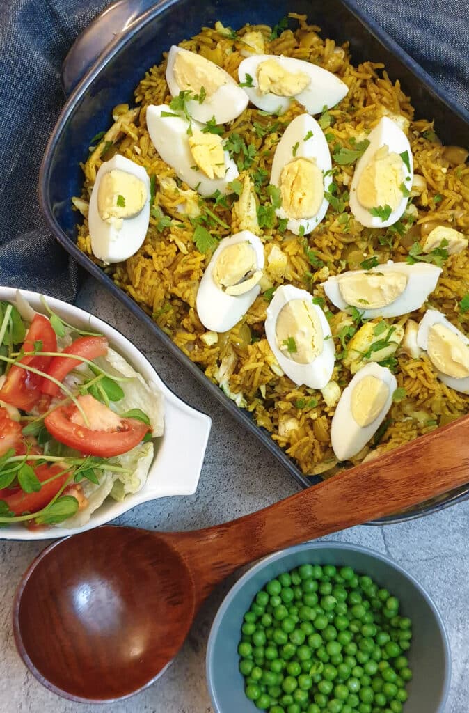 Smoked haddock kedgeree in a blue serving dish next to a tomato salad and a bowl of peas, with a wooden serving spoon.