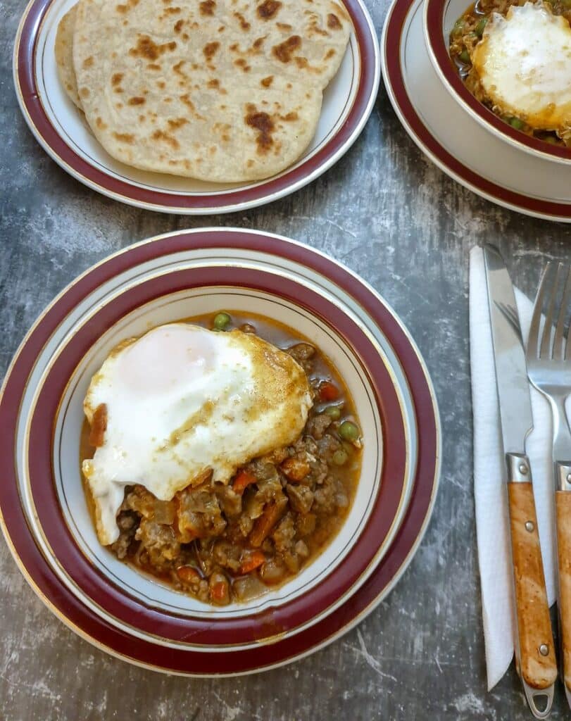 Lamb keema topped with a baked egg in a dish next to a plate of flatbreads.