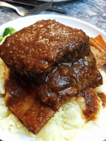 A piece of short rib on a pile of mashed potatoes on a white plate with vegetables and gravy.
