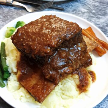 A piece of short rib on a pile of mashed potatoes on a white plate with vegetables and gravy.