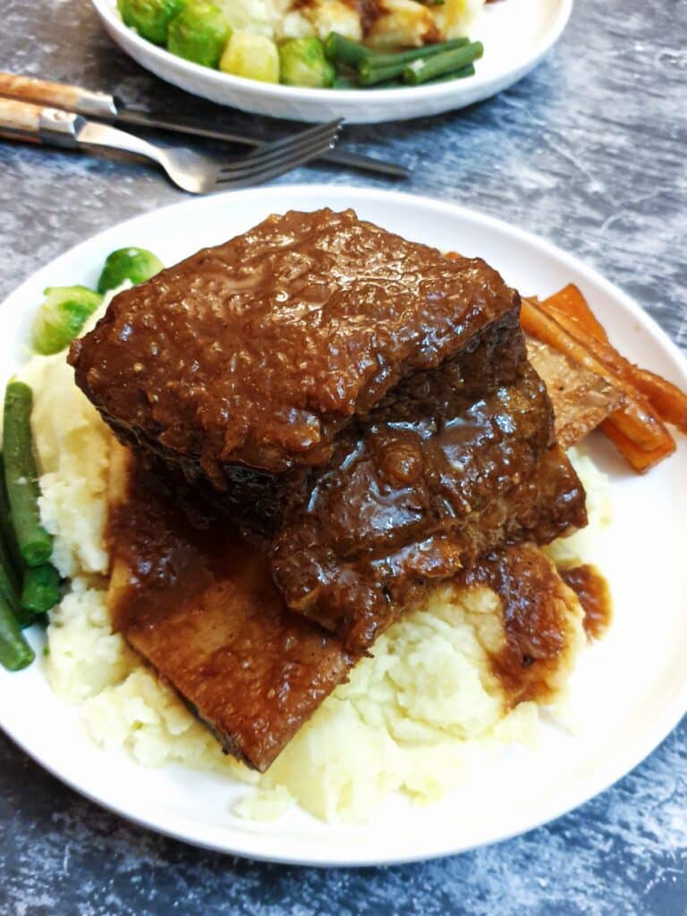 A piece of short rib on a pile of mashed potatoes on a white plate with vegetables and gravy.