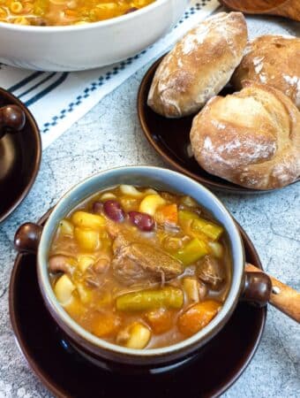 A dish of beef minestrone soup next to a plate of crusty bread rolls.