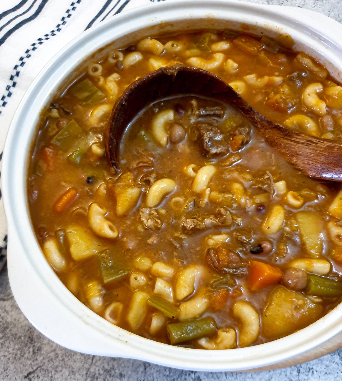 Beef minestrone soup in a white serving dish with a wooden serving spoon.