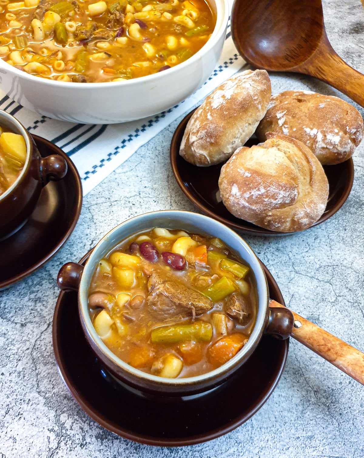 A dish of beef minestrone soup next to a plate of crusty bread rolls.