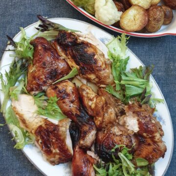 Cooked spatchcock chicken pieces on a serving plate next to a dish of vegetables.
