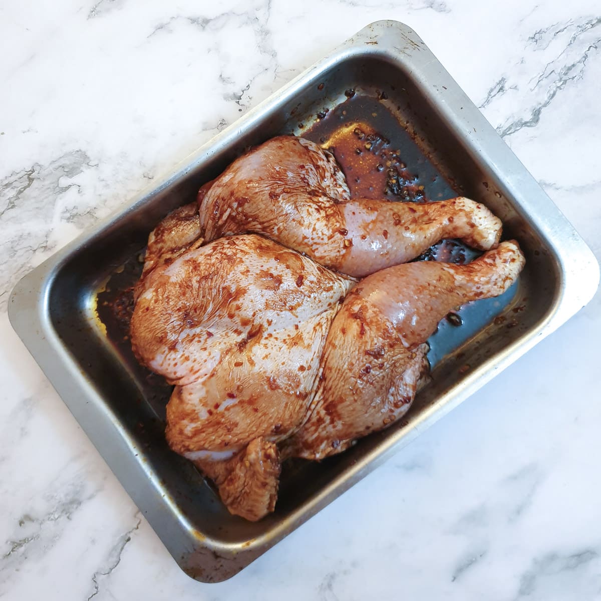 A spatchcock chicken covered in honey, chilli and garlic marinade, in a roasting pan.