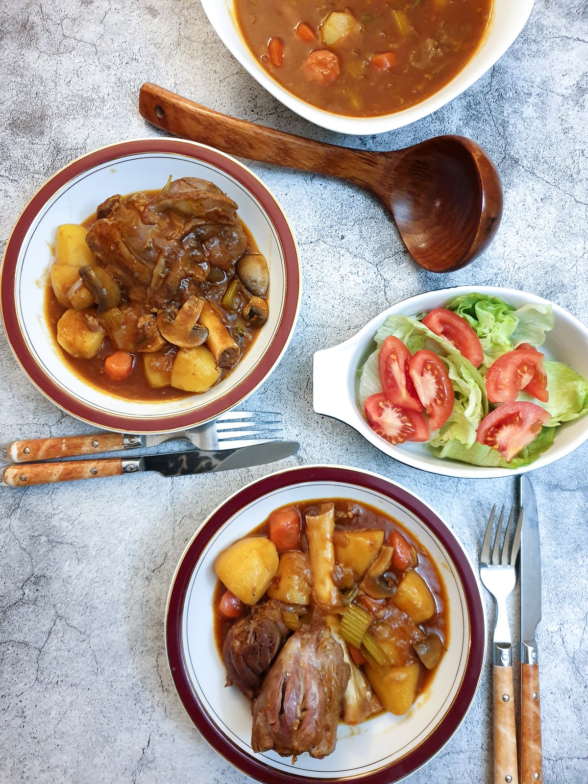 Two dishes of lamb shanks on a table alongside a greed salad and a dish of vegetables in gravy.