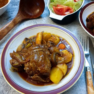 A lamb shank in a dish surrounded by vegetables and thick tomato gravy.