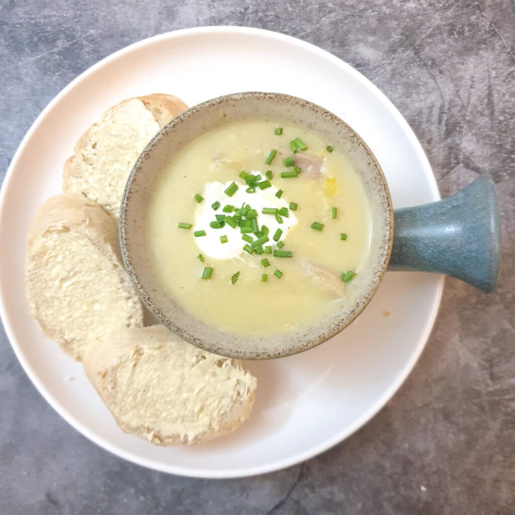 A soup dish filled with potato and leek soup on a while place with slices of crusty bread.