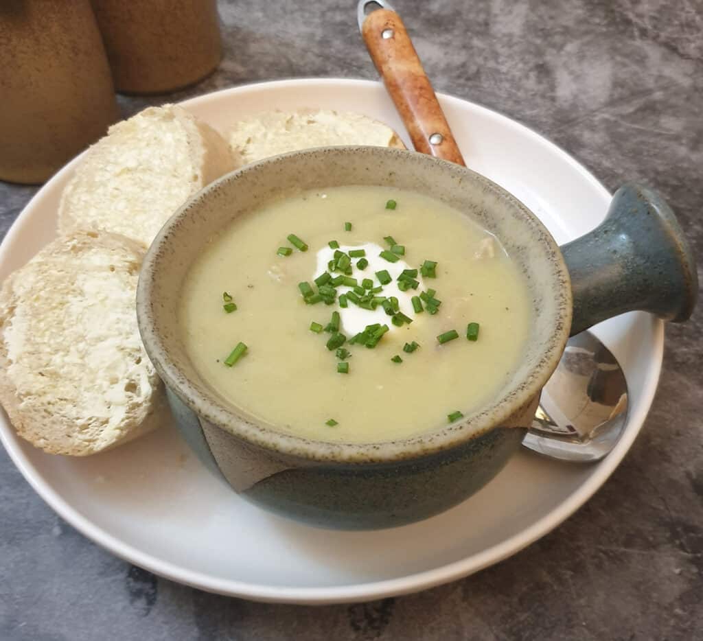 A soup dish filled with potato and leek soup on a while place with slices of crusty bread.