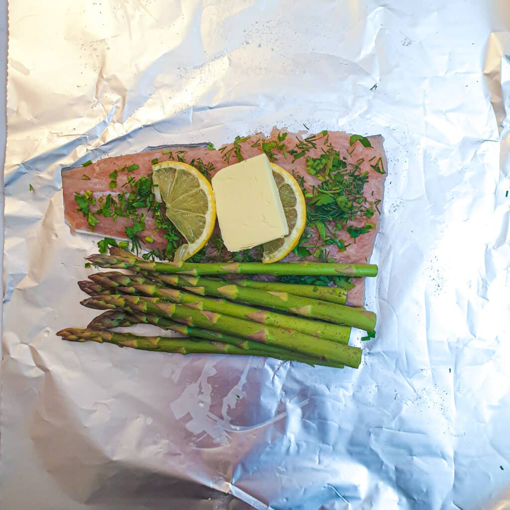 A piece of uncooked salmon topped with herbs, lemon and a knob of butter on a sheet of tin foil.