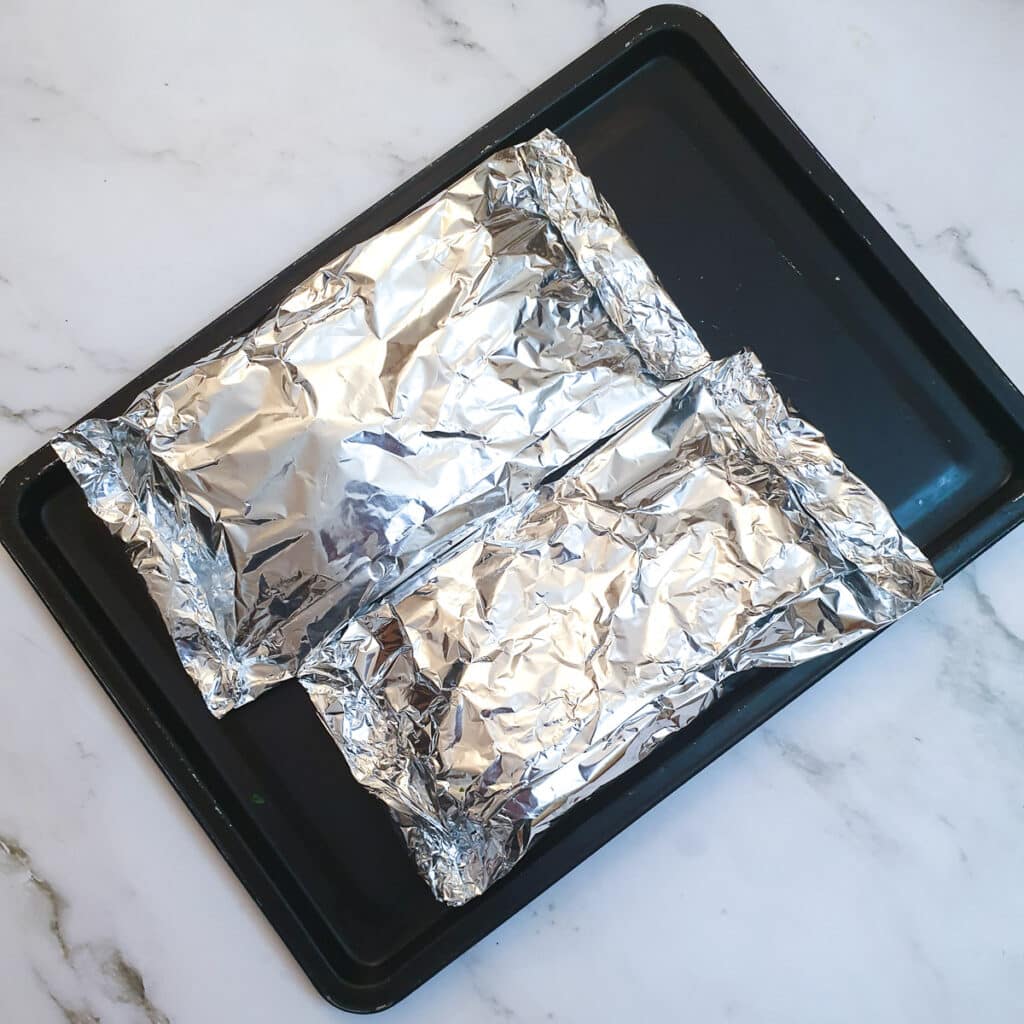 Two tin foil parcels of salmon and asparagus on a baking tray.