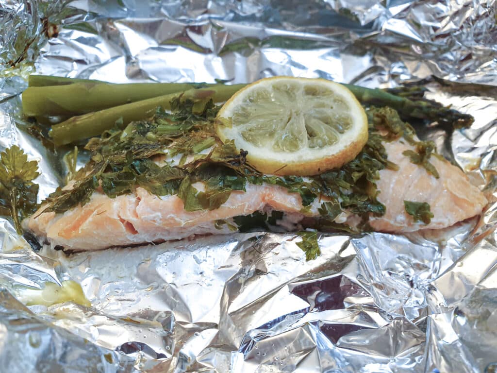 Close up of a piece of cooked salmon on a sheet of tin foil, covered with herbs and lemon, with asparagus spears.