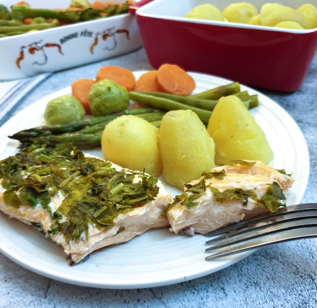 A piece of salmon covered in dill and parsley on a plate with potatoes and vegetables.