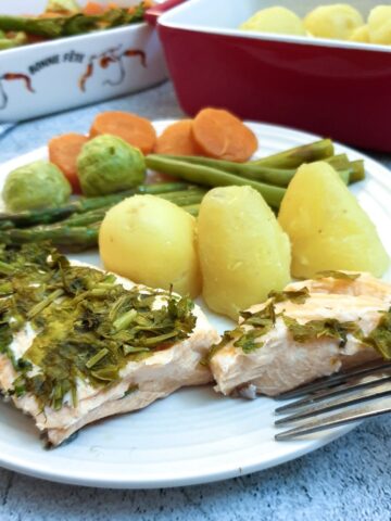 A piece of salmon covered in dill and parsley on a plate with potatoes and vegetables.