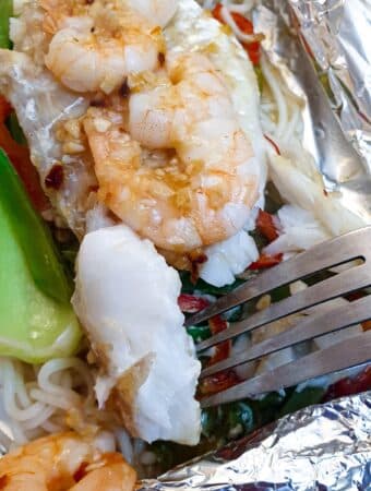 Close up of a piece of cooked fish on a pork held above a dish of cod and prawn fish parcels.
