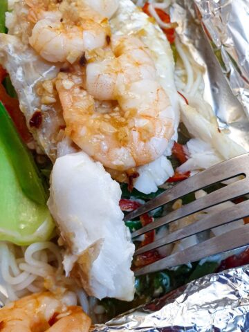 Close up of a piece of cooked fish on a pork held above a dish of cod and prawn fish parcels.