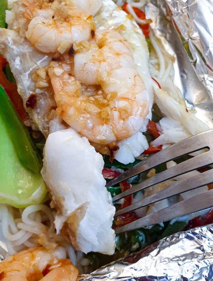 Close up of a piece of cooked fish on a pork held above a dish of cod and prawn fish parcels.
