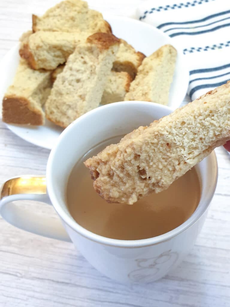 A buttermilk rusk being dunked into a cup of coffee with a plate of rusks in the background.