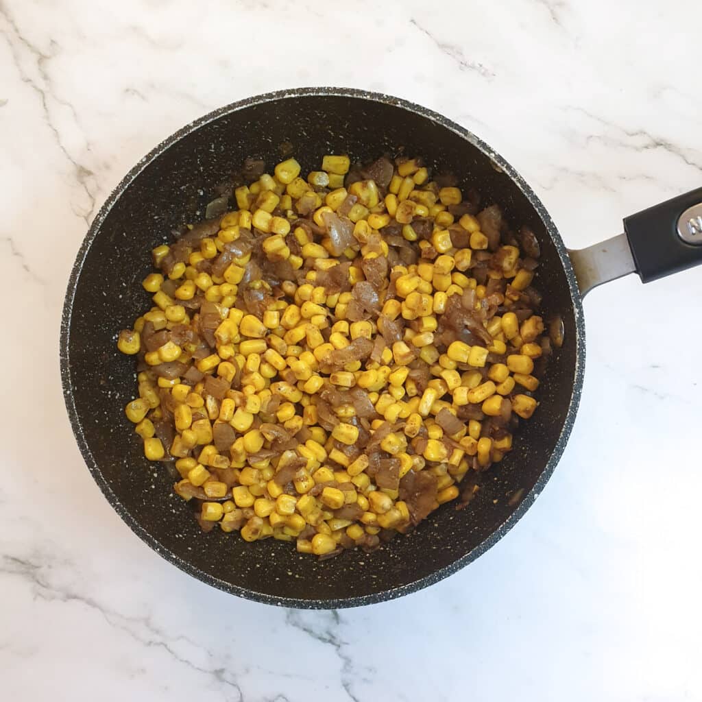 Canned sweetcorn mixed with onions and spices in a frying pan.