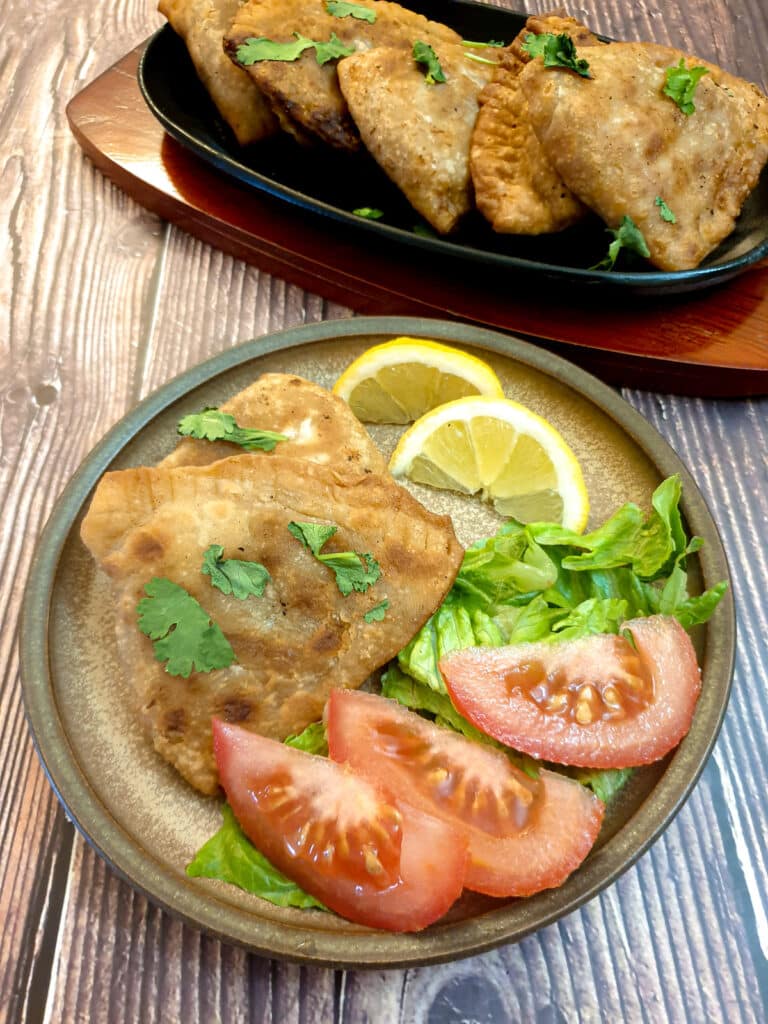 2 samosas on a brown plate with lettuce, slices of tomato  and lemon wedges.