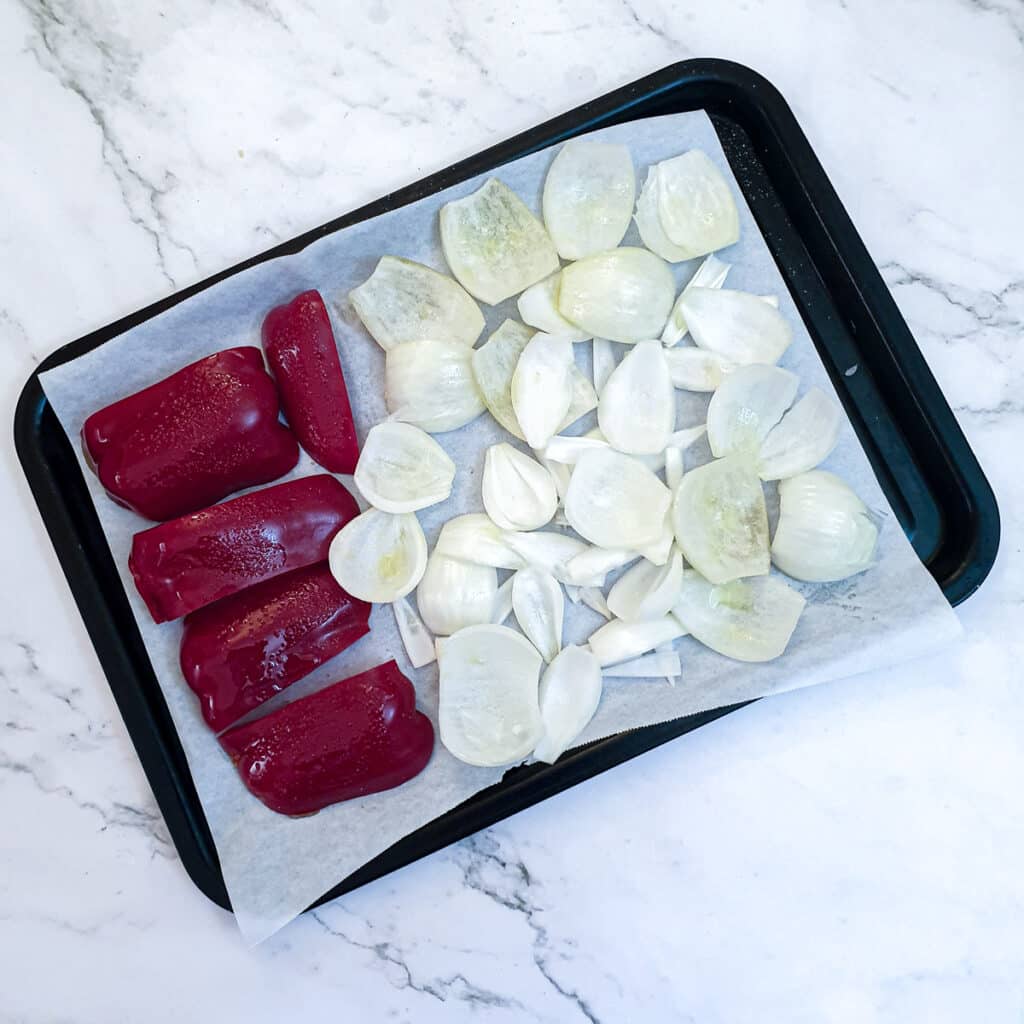 Uncooked pieces of red pepper and onions on a baking tray.