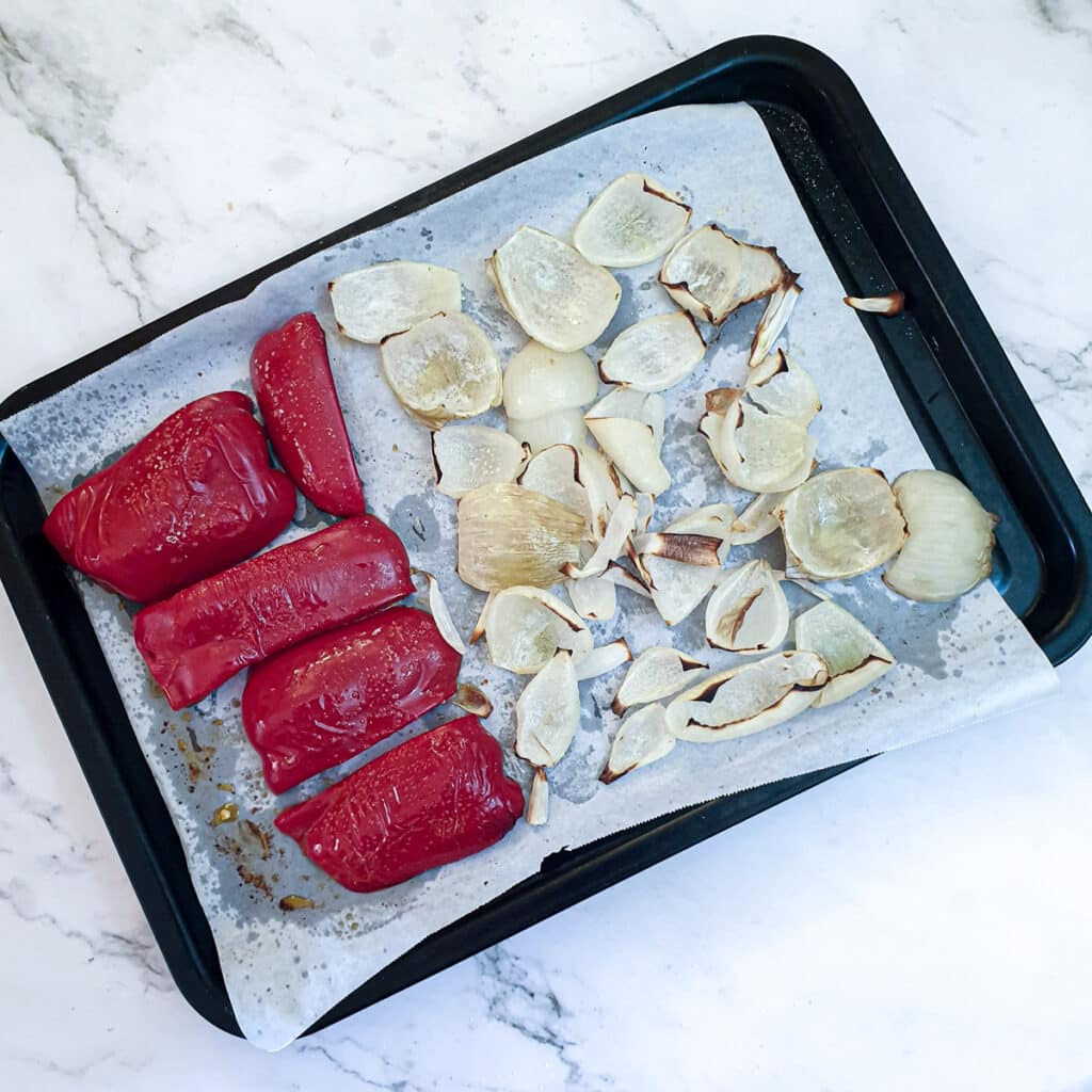 Cooked pieces of red pepper and onions on a baking tray.