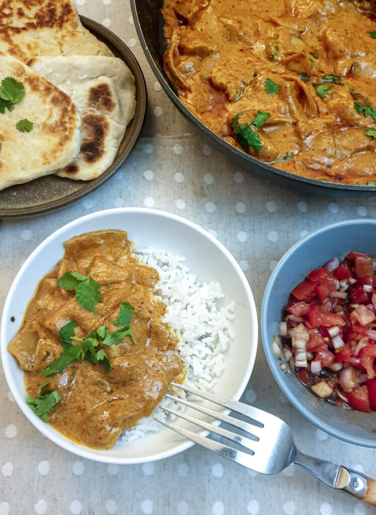 A white bowl containing rice and butter chicken alongside dishes containing tomato sambal and garlic naan bread.