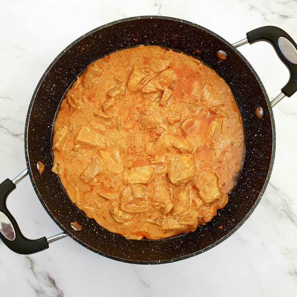 A frying pan containing pieces of chicken breast in a buttery tomato curry sauce.