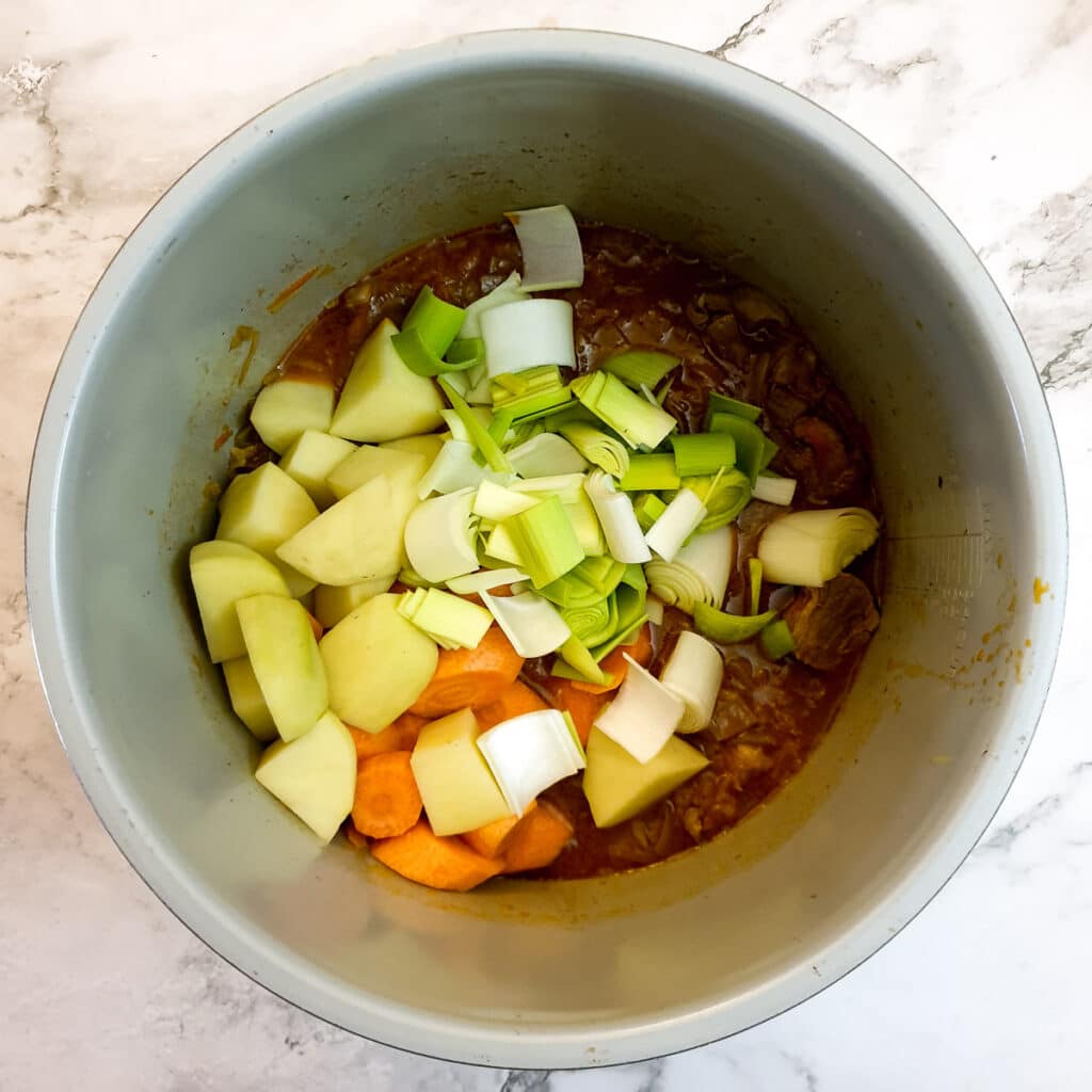 Potatoes, carrots and leeks being added to the cooked meat in a pressure cooker.